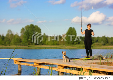 Woman with a fishing rod in hand and small Yorkshire Terrier dog standing on wooden bridge, pier, footbridge on a river, lake at summer day. Nature recreation. Beautiful landscape. Blurred photography Woman with a fishing rod in hand and small Yorkshire Terrier dog standing on wooden bridge, pier, footbridge on a river, lake at summer day. Nature recreation. Beautiful landscape. Blurred photography 85925397