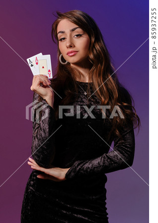 Brunette woman in black velvet dress showing two playing cards, posing against coloful background. Gambling entertainment, poker, casino. Close-up. Brunette woman in black velvet dress showing two playing cards, posing against coloful background. Gambling entertainment, poker, casino. Close-up. 85937255