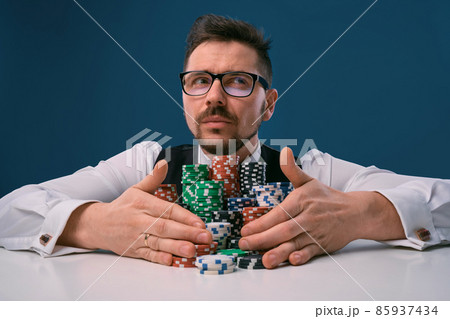 Guy in glasses, black vest and shirt sitting at white table with stacks of chips on it, posing on blue background. Gambling, poker, casino. Close-up. Guy in glasses, black vest and shirt sitting at white table with stacks of chips on it, posing on blue background. Gambling, poker, casino. Close-up. 85937434