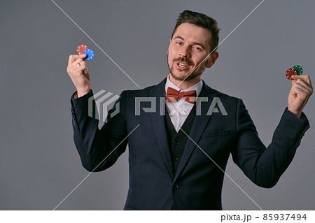 Man in black classic suit and red bow-tie showing some colored chips, posing against gray studio background. Gambling, poker, casino. Close-up. 85937494