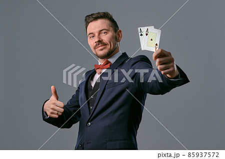 Man in black classic suit and red bow-tie showing two playing cards while posing against gray studio background. Gambling, poker, casino. Close-up. 85937572
