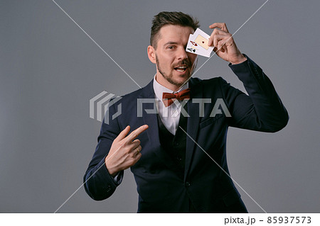 Man in black classic suit and red bow-tie showing two playing cards while posing against gray studio background. Gambling, poker, casino. Close-up. Man in black classic suit and red bow-tie showing two playing cards while posing against gray studio background. Gambling, poker, casino. Close-up. 85937573