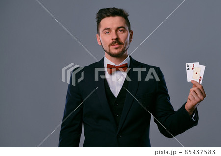 Man in black classic suit and red bow-tie showing two playing cards while posing against gray studio background. Gambling, poker, casino. Close-up. 85937583