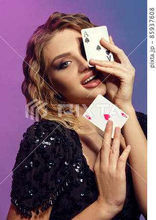 Brown-haired female in black shiny dress is smiling, showing two playing cards, posing on colorful background. Gambling, poker, casino. Close-up. 85937608