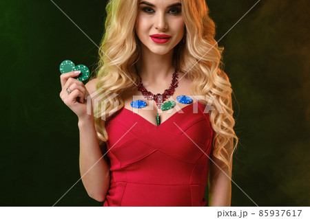 Blonde female in red dress. Smiling, showing two green chips and some colorful on her decollete. Colorful smoky background. Poker, casino. Close-up Blonde female in red dress. Smiling, showing two green chips and some colorful on her decollete. Colorful smoky background. Poker, casino. Close-up 85937617