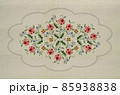 Embroidery ornament of twigs with flowers of pink-red roses with poppies and blue flowers with a wavy oval gray frame on a background of beige fabric. 85938838