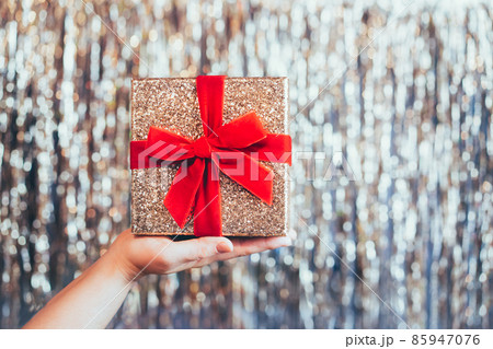Female hand holding glittering golden gift box with velvet red ribbon with a bow on the sparkling gold tinsel background. The concept of a surprise gift for birthday, Xmas. Festive background. Female hand holding glittering golden gift box with velvet red ribbon with a bow on the sparkling gold tinsel background. The concept of a surprise gift for birthday, Xmas. Festive background. 85947076