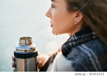 Young pretty woman holds a thermos and drinks tea wrapped in blanket over blur river background. 85951423