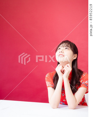Beautiful asian woman wearing traditional cheongsam qipao dress with hands to her chin, looking to the side on red background. Beautiful asian woman wearing traditional cheongsam qipao dress with hands to her chin, looking to the side on red background. 85952219