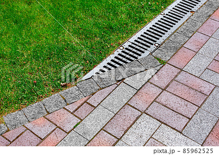 drainage grate bolted to storm drain at corner of pedestrian pavement walk way made of stone brick tiles gray and red with pattern in backyard with green grass on sunny summer park, nobody. 85962525