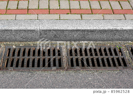 catch basin grate of the lattice of the drain system for drainage of rainwater from the road surface with a tarmac road at the curb with a stone brick tile sidewalk close-up, nobody. catch basin grate of the lattice of the drain system for drainage of rainwater from the road surface with a tarmac road at the curb with a stone brick tile sidewalk close-up, nobody. 85962528