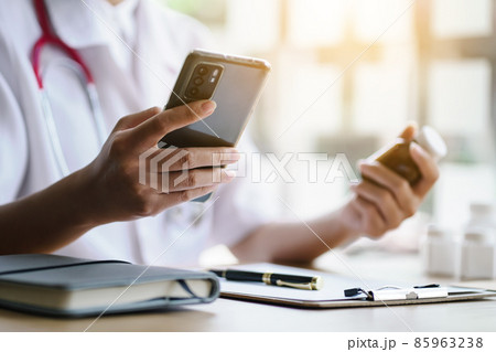 Female doctor holding a cell phone and a bottle of medicine to check the expiration date in the online pharmaceutical register. Female doctor holding a cell phone and a bottle of medicine to check the expiration date in the online pharmaceutical register. 85963238