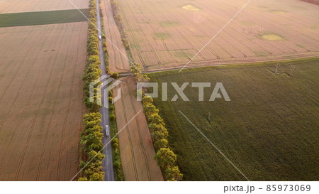 Drone flying over road between wheat fields during dawn sunset Drone flying over road between wheat fields during dawn sunset 85973069