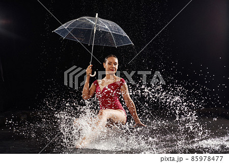 Beautiful young girl in red sportswear with umbrella in aqua studio. Drops of water spread about her fitness body. The perfect figure on the background of water splashes. Classic dark color background 85978477