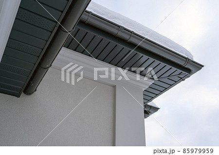 Corner of the house with new gray metal tile roof and rain gutter covered by snow at winter. Metallic Guttering System, Guttering and Drainage Pipe Exterior 85979959
