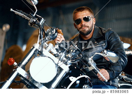 Cool bearded man biker in sunglasses sitting on his motorbike and smoking cigar Cool bearded man biker in sunglasses sitting on his motorbike and smoking cigar 85983065