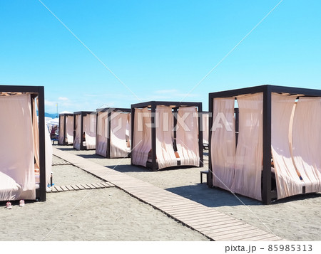 Beach cabins with a metal frame and light curtains stand in a row on the sand with wooden paths between them Beach cabins with a metal frame and light curtains stand in a row on the sand with wooden paths between them 85985313