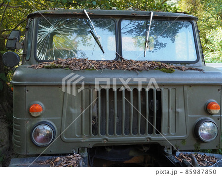 An old abandoned military truck with broken glass stands strewn with dry foliage surrounded by green trees An old abandoned military truck with broken glass stands strewn with dry foliage surrounded by green trees 85987850