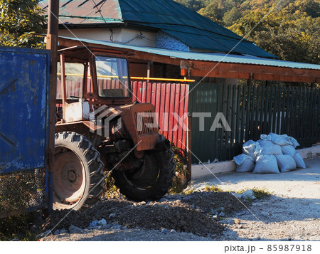 An old red tractor stands on a broken rocky road near an old fenced private house 85987918
