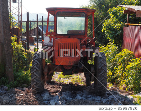 An old red tractor stands on a broken rocky road in a village on a summer sunny day 85987920