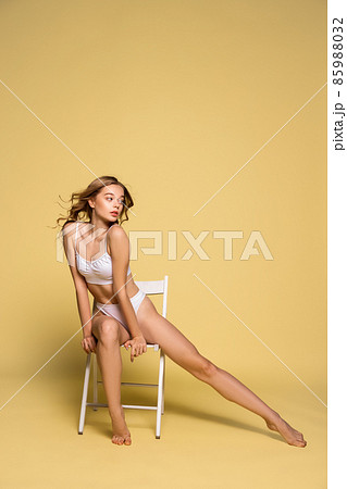 Studio shot of young beautiful pretty girl in underwear sitting on wooden chair isolated over yellow background. Natural beauty of female body. Studio shot of young beautiful pretty girl in underwear sitting on wooden chair isolated over yellow background. Natural beauty of female body. 85988032