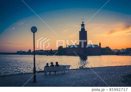 Watercolor drawing of Silhouette of women sitting on bench at promenade of Lake Malaren looking at Stockholm City Hall Stadshuset tower 85992717