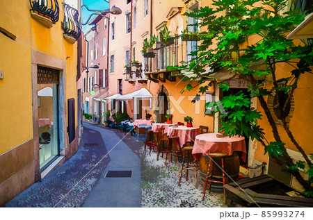 Watercolor drawing of restaurant tables on wooden barrels and chairs in typical italian narrow street and traditional colorful buildings 85993274