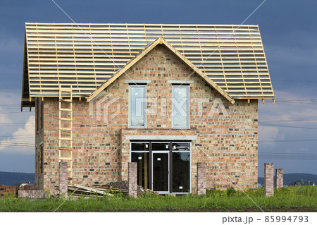 Two-story residential house with plastic windows, wide door and wooden frame for roof under construction. High wooden ladder at brick walls and brick columns for fence on dark blue sky background. Two-story residential house with plastic windows, wide door and wooden frame for roof under construction. High wooden ladder at brick walls and brick columns for fence on dark blue sky background. 85994793