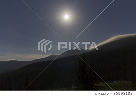 Night view of bright moon shining in dark blue sky over mountains covered with evergreen forest and small tourist tent on grassy valley. Beauty and magic of nature, tourism and traveling concept. 85995105