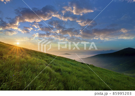 Mountain landscape in nice weather at sunrise. Green grassy steep hill, foggy valley and distant mountains under bright blue sky with lit by raising sun white clouds. Beauty of nature concept. 85995203