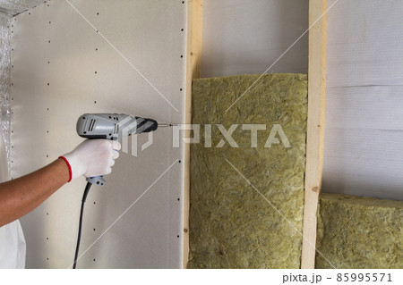 Close-up of worker hand with electric screwdriver fastening drywall to wooden timber frame with insulation mineral rock wool staff. Warm home, low energy bills, construction and renovation concept. Close-up of worker hand with electric screwdriver fastening drywall to wooden timber frame with insulation mineral rock wool staff. Warm home, low energy bills, construction and renovation concept. 85995571