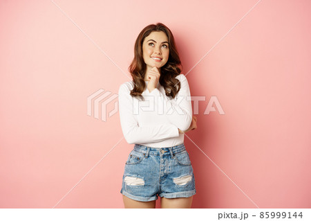 Beautiful romantic girlfriend, looking at upper left corner and smiling thoughtful, thinking, having an idea, standing over pink background in stylish clothes 85999144