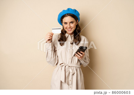 Portrait of trendy woman in coat holding smartphone, showing credit card, shopping online, order something on mobile phone, standing over beige background Portrait of trendy woman in coat holding smartphone, showing credit card, shopping online, order something on mobile phone, standing over beige background 86000603