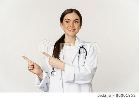 Portrait of smiling young woman doctor, healthcare medical worker, pointing fingers left, showing clinic promo, logo or banner, standing over white background 86001855