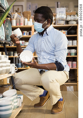 Man in protective mask carefully chooses ceramic mugs in tableware store Man in protective mask carefully chooses ceramic mugs in tableware store 86002068