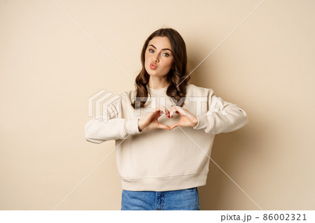 Beautiful, romantic brunette woman showing heart, love sign, care for you, smiling at camera, standing over beige background Beautiful, romantic brunette woman showing heart, love sign, care for you, smiling at camera, standing over beige background 86002321