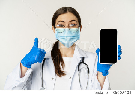 Online medical help concept. Enthusiastic young woman doctor in face mask, showing thumbs up and mobile phone app, smartphone screen interface, white background Online medical help concept. Enthusiastic young woman doctor in face mask, showing thumbs up and mobile phone app, smartphone screen interface, white background 86002401