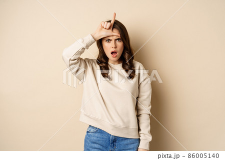 Woman showing loser sign on forehead, L word, standing over beige background Woman showing loser sign on forehead, L word, standing over beige background 86005140
