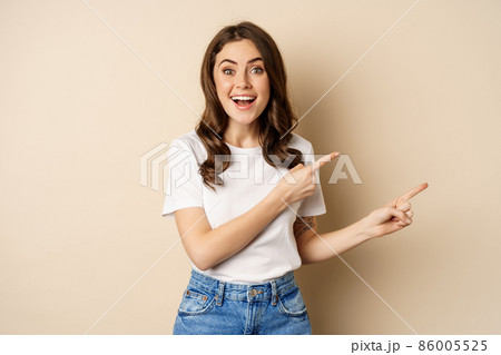 Smiling brunette woman in t-shirt, pointing fingers right, showing promo offer or advertisement, demonstrating banner, standing against beige background 86005525