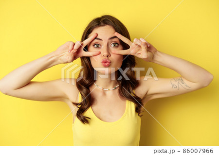 Close up portrait of smiling beautiful girl, showing peace v-sign and looking happy, posing for summer photo, standing against yellow background Close up portrait of smiling beautiful girl, showing peace v-sign and looking happy, posing for summer photo, standing against yellow background 86007966