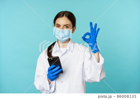 Online doctor and clinic. Young woman in medical face mask, using smartphone for client remote online appointment, showing okay sign, standing over blue background 86008135