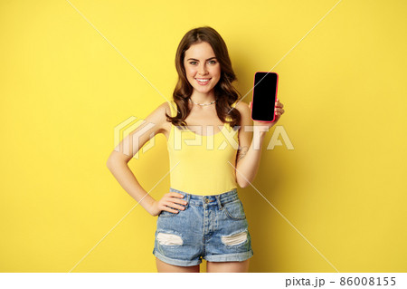 Stylish smiling woman showing mobile phone screen, app interface on smartphone, standing in tank top and shorts against yellow background 86008155