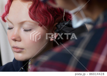 A beautician makes a puncture of the earlobe with a disposable device in a beauty salon. Ear piercing lobe. 86014428