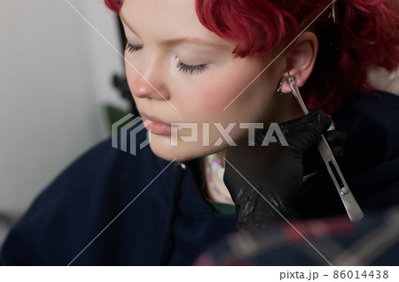 A beautician makes a puncture of the earlobe with a disposable device in a beauty salon. Ear piercing lobe. A beautician makes a puncture of the earlobe with a disposable device in a beauty salon. Ear piercing lobe. 86014438
