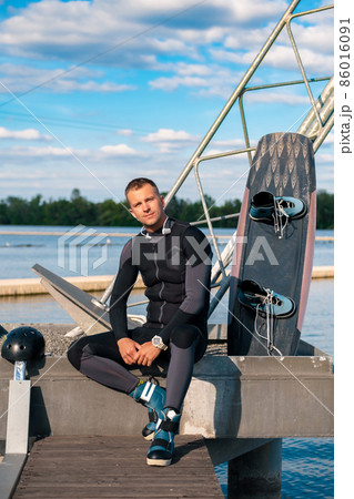 Relaxed sporty man in wetsuit sitting on pier with wakeboard 86016091