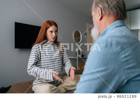 Pretty young woman and mature male having fun, enjoying leisure time, sharing news at home. Back view of unrecognizable bearded grandfather talking with pretty young granddaughter sitting in bed. Pretty young woman and mature male having fun, enjoying leisure time, sharing news at home. Back view of unrecognizable bearded grandfather talking with pretty young granddaughter sitting in bed. 86021289