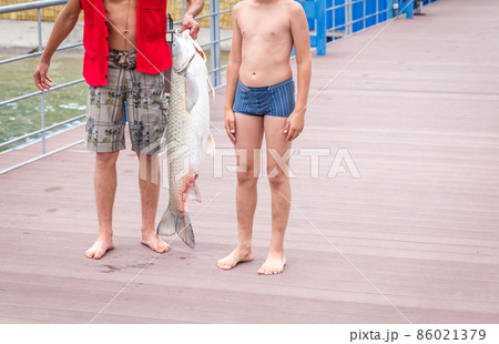 Fisherman holding a huge grass carp on a pier on a lake, standing with his son Fisherman holding a huge grass carp on a pier on a lake, standing with his son 86021379