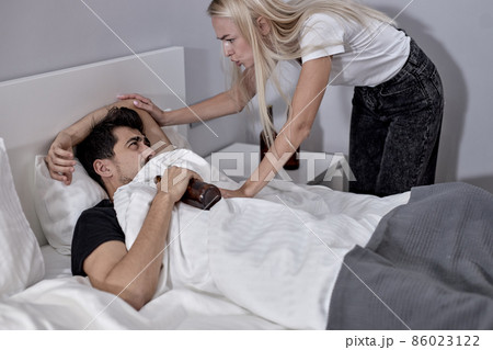 Alcoholic male lying on bed with alcohol while irritated young wife is scolding him 86023122