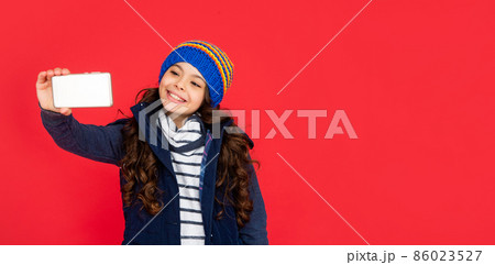 happy kid making selfie in hat on red background. teen girl in down vest with smartphone. 86023527