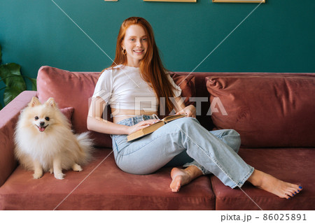 Portrait of smiling young woman reading book sitting on cozy sofa with pretty spitz pet dog at apartment with modern interior, looking at camera. Happy female spending pleasant weekend with doggy. 86025891